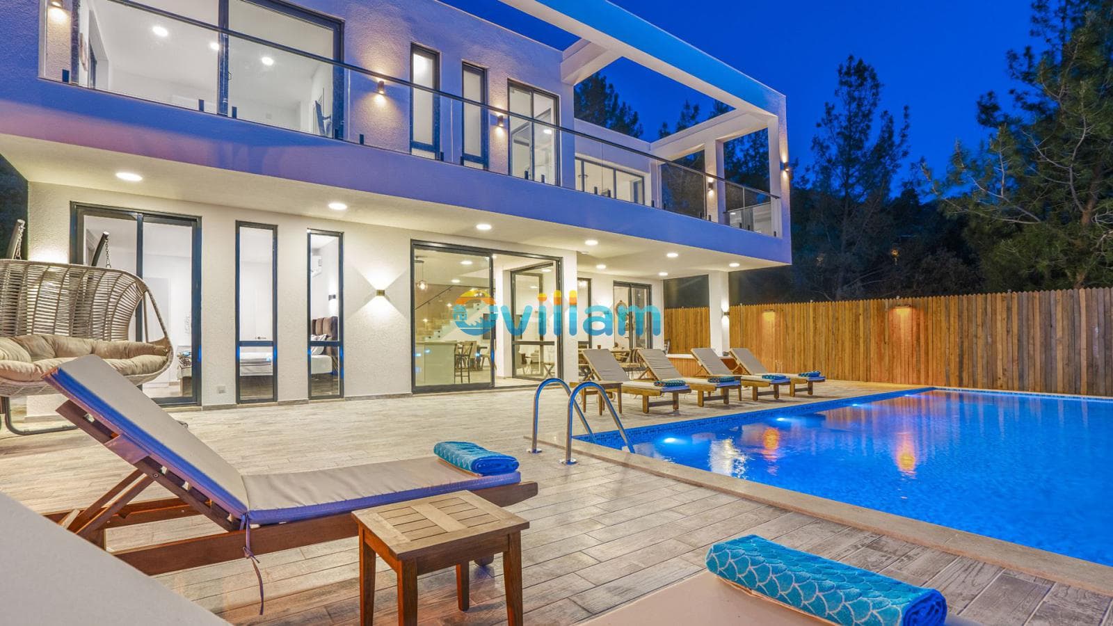 Akşam aydınlatmalı modern villanın önünde havuz, şezlonglar ve asılı bir salıncak.