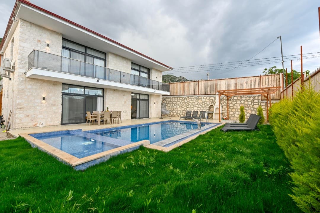 Taş kaplama iki katlı villa, önünde yüzme havuzu, çim bahçe, şezlonglar ve yemek masası bulunuyor.