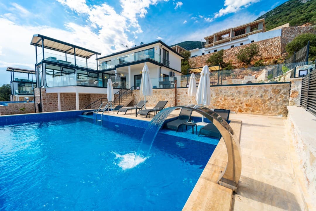 Taş duvarlı terasta şelale özellikli yüzme havuzu, şezlonglar ve güneş şemsiyeleri bulunan modern villa.