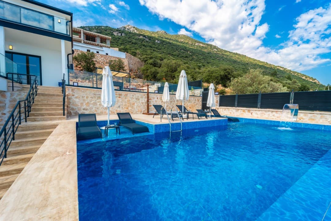 Dağ manzaralı havuz başında şezlonglar ve beyaz şemsiyeler bulunan villa.