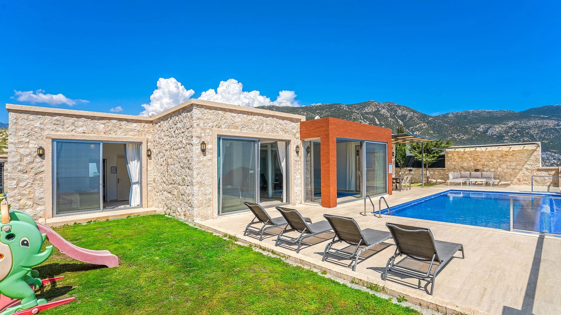 Taş duvarlı villa, önünde havuz, şezlonglar ve çocuk kaydırağı bulunan güneşli bir bahçe.