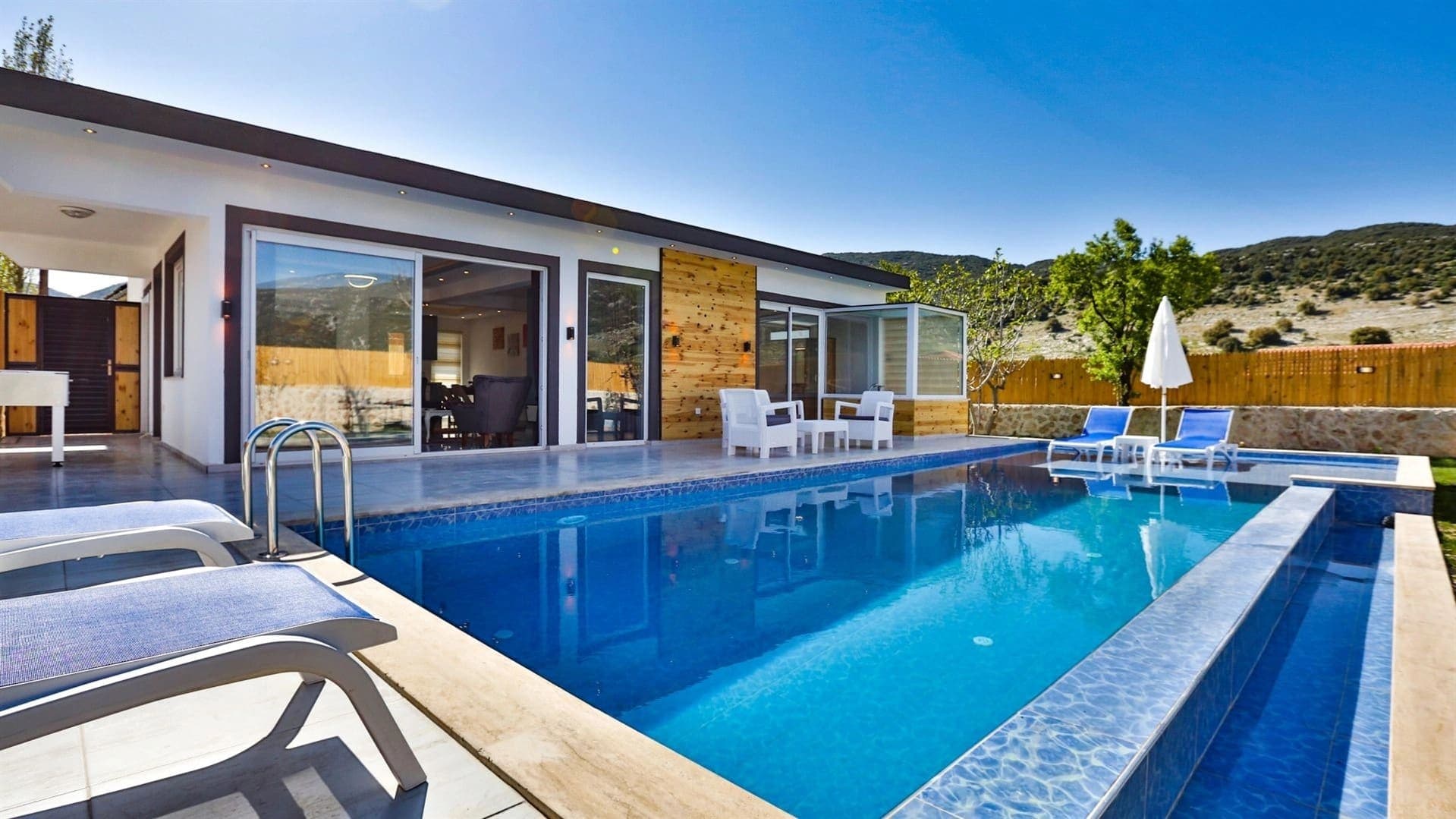 Önünde havuzu, şezlongları ve güneş şemsiyesi bulunan modern dış cepheli tek katlı villa.