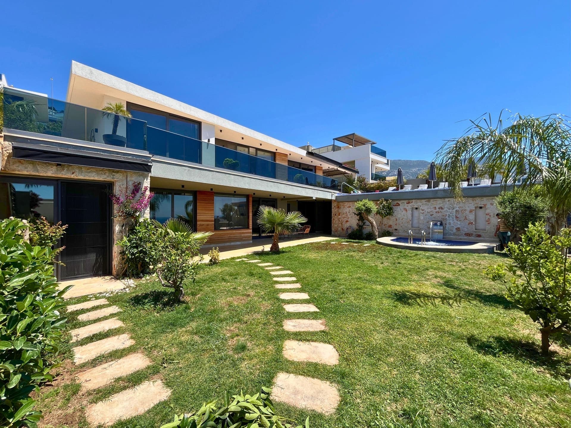 Yeşil bahçeli, taş duvarlı ve modern balkonlu villa dış cephesi.