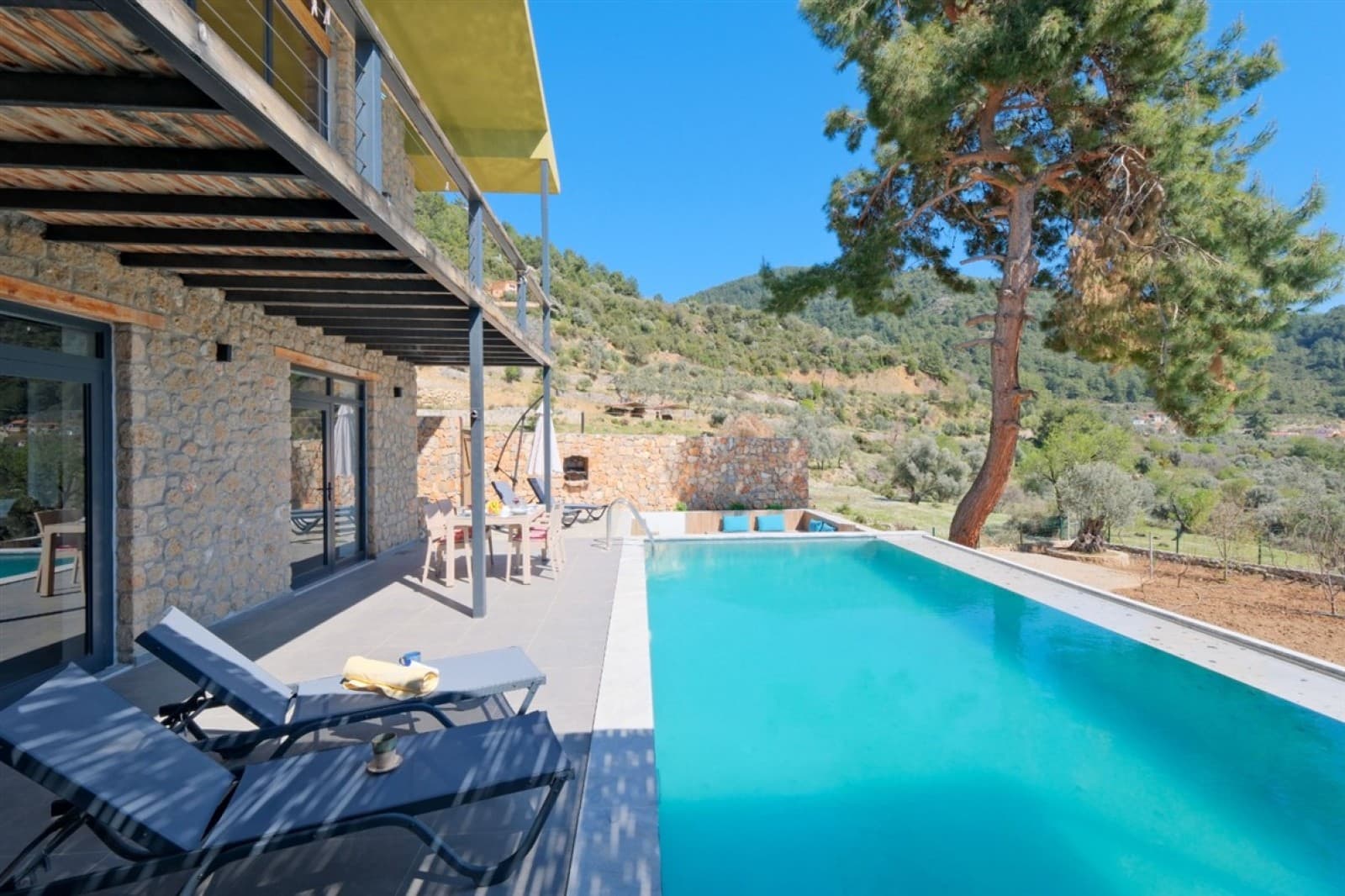 Çam ağaçları ve dağ manzaralı havuz kenarında şezlonglar.