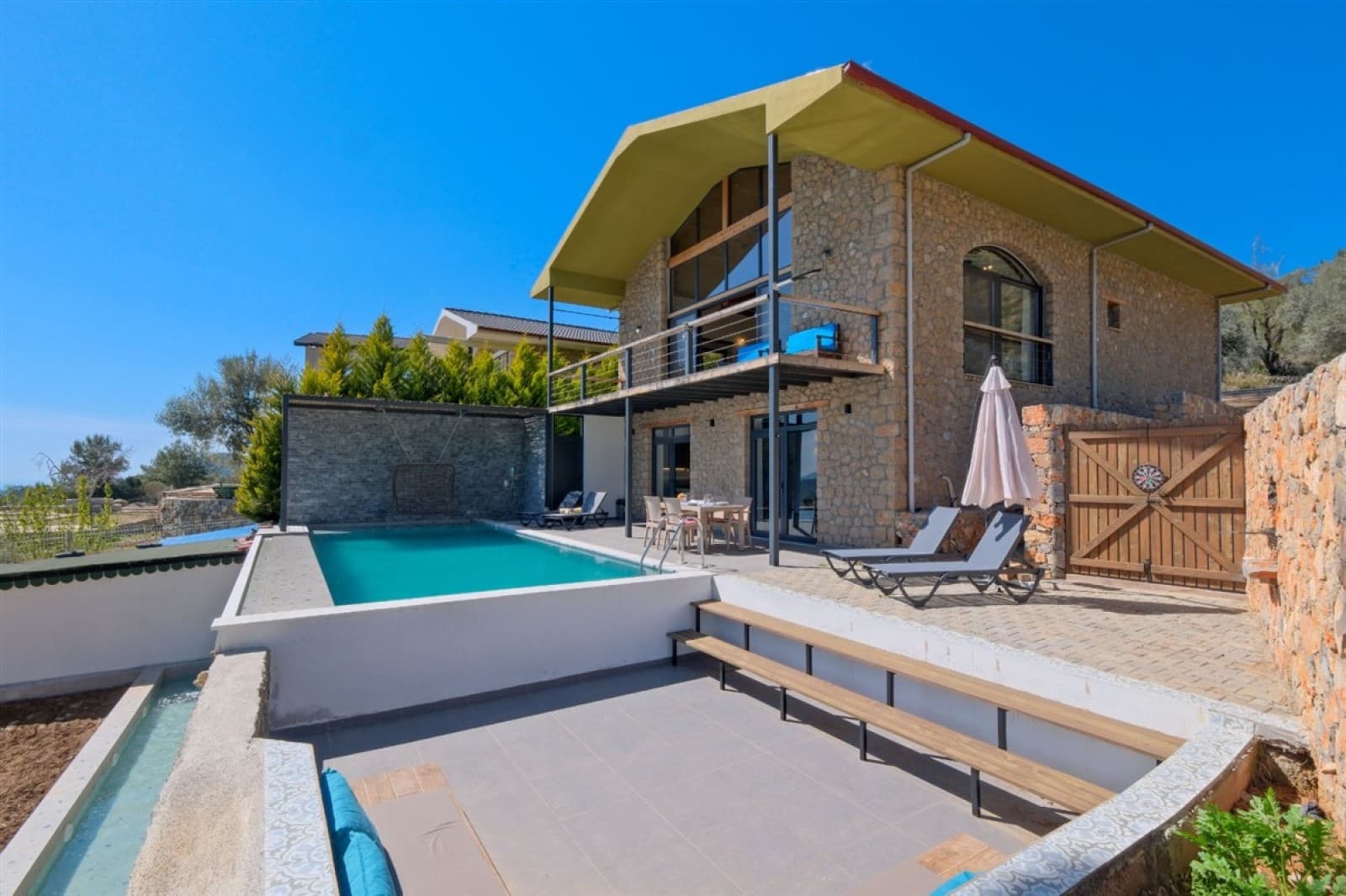 Taş kaplama cepheli, balkonlu ve önünde havuz bulunan iki katlı villa.