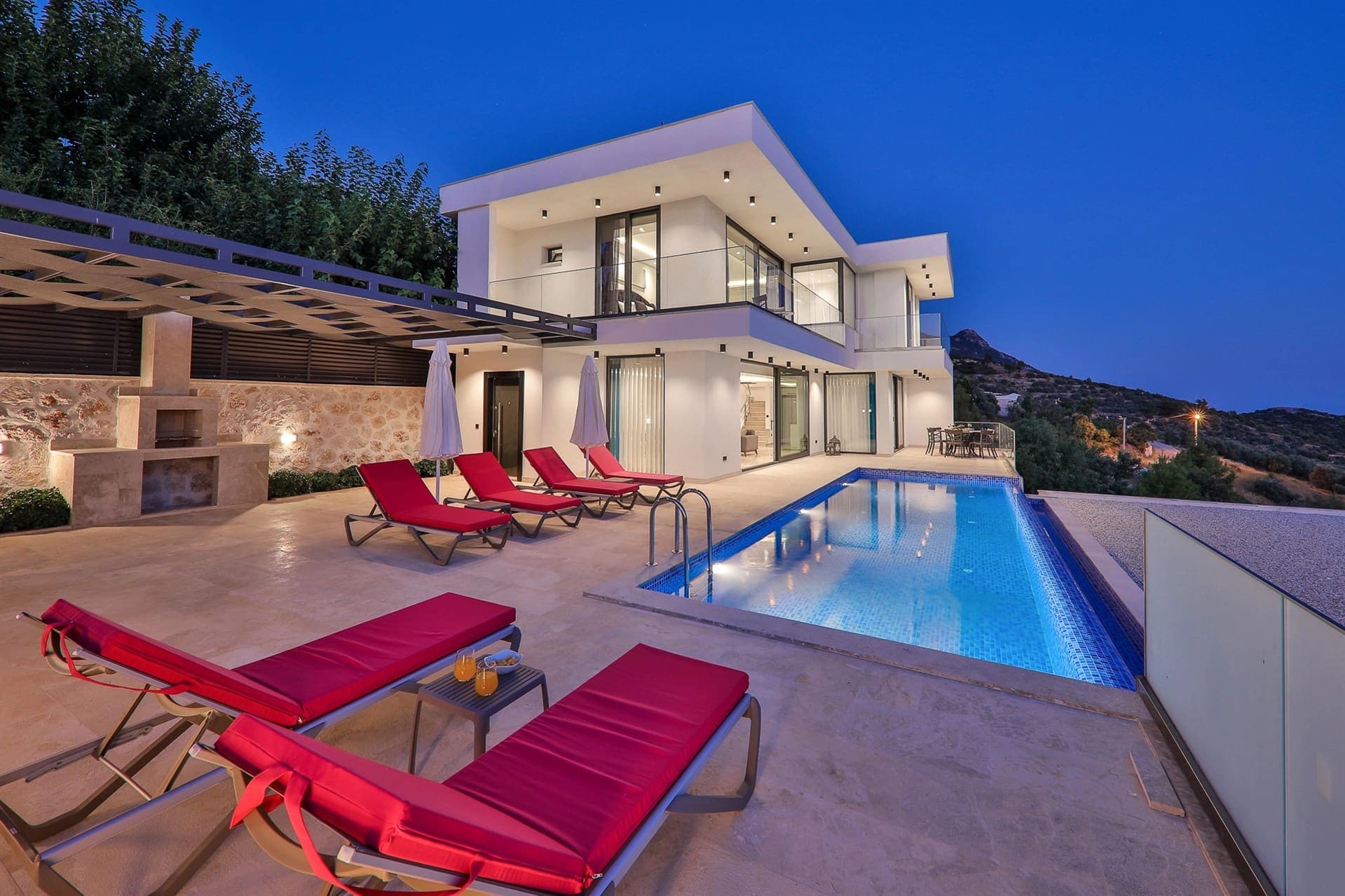 Akşam ışıklandırmalı modern villa, özel havuz, kırmızı şezlonglar ve taş barbekü alanı.