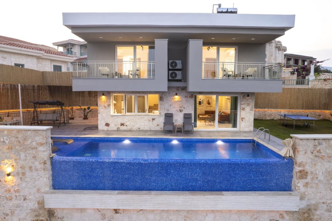 Önünde mavi havuz, bahçesinde şezlonglar ve salıncak olan iki katlı gri villa.