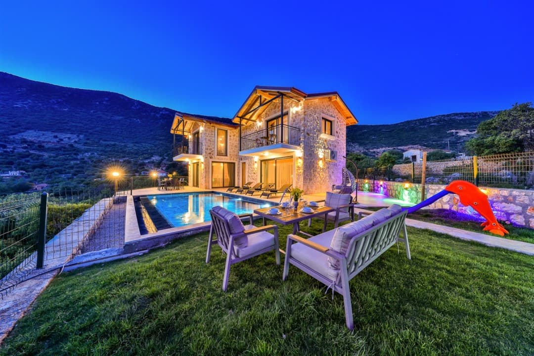 Akşam vakti aydınlatılmış taş villa, havuz, bahçe oturma grubu ve dağ manzarası.