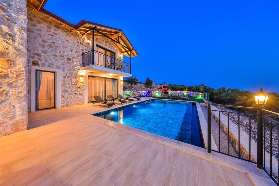 Akşam vakti aydınlatılmış taş villa, havuz başındaki şezlonglar ve bahçe manzarası.