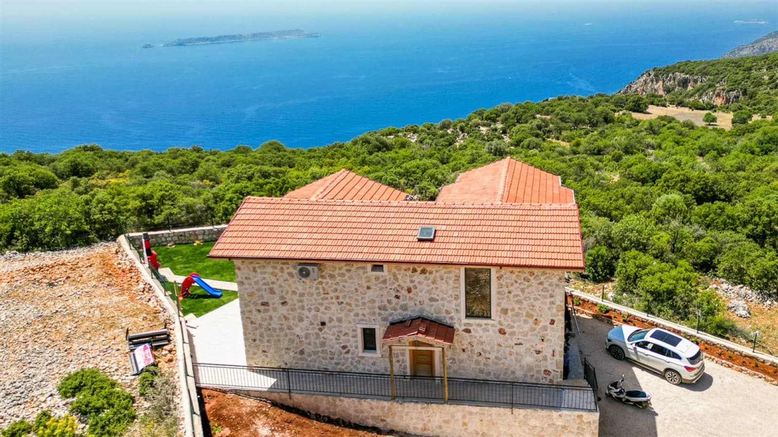 Ağaçlar arasında taş cepheli villa ve uzakta deniz manzarası.