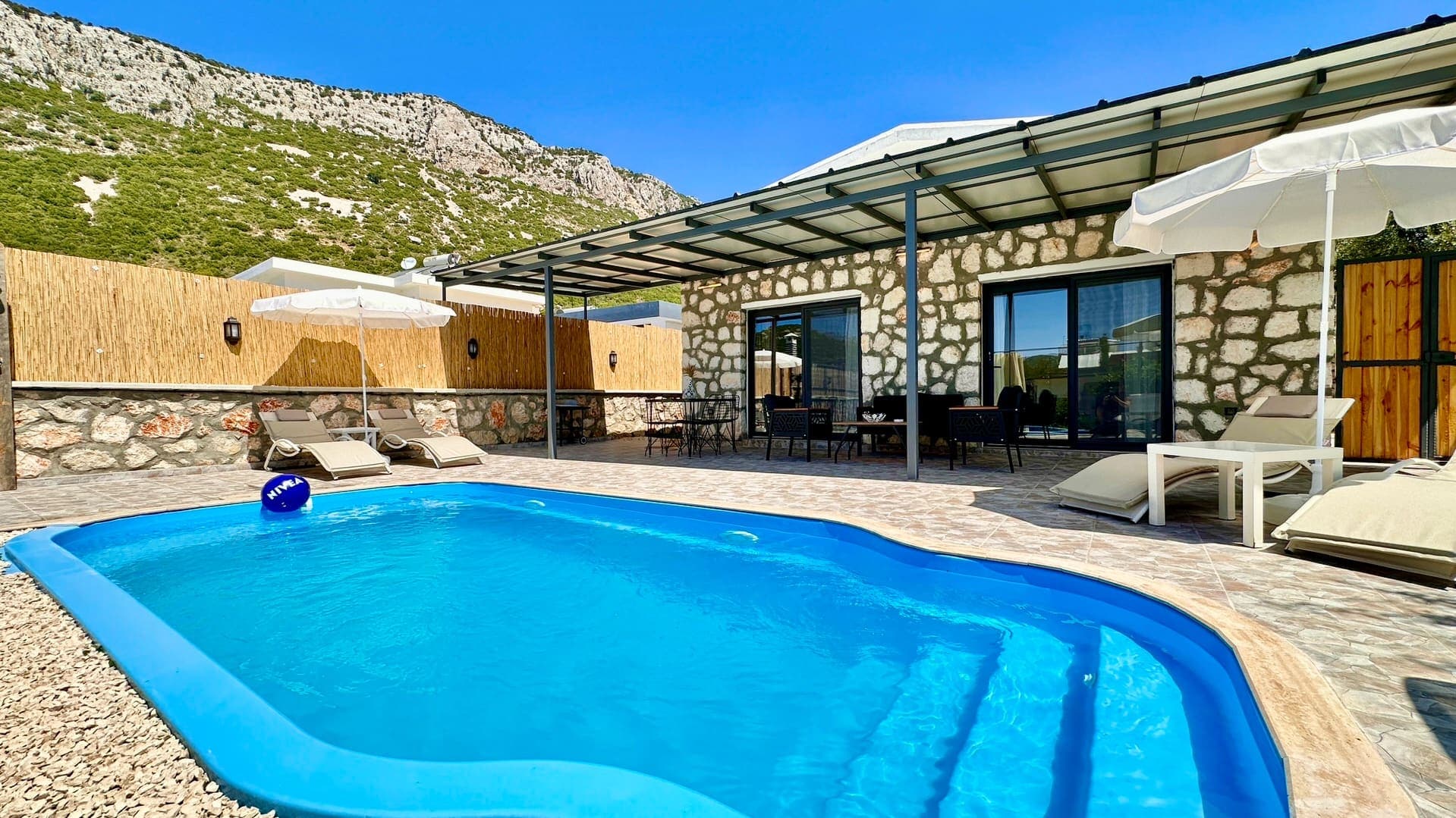 Taş duvarlı villa, yüzme havuzu, şezlonglar ve bahçe mobilyaları.