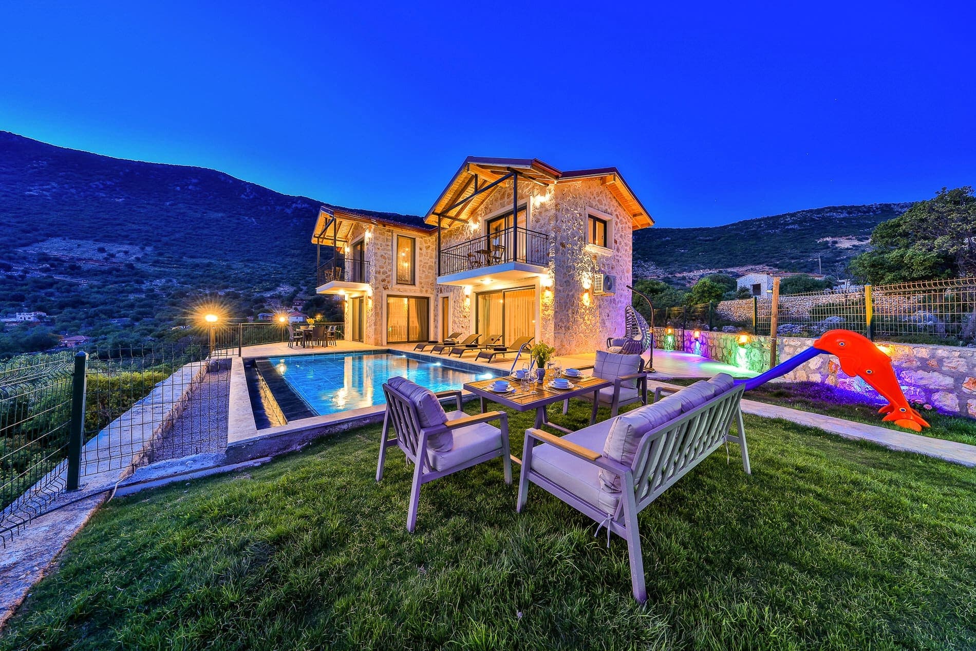 Gece aydınlatmalı taş villa, havuz, bahçe mobilyaları ve çocuk kaydırağı.