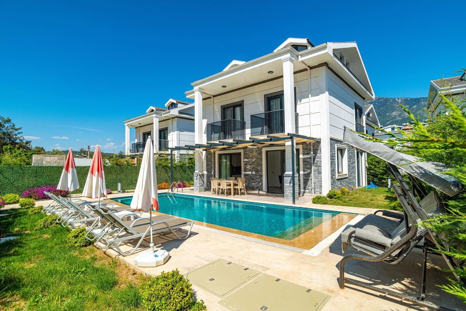 Bahçesinde havuz, şezlonglar ve salıncak bulunan beyaz dış cepheli villa.