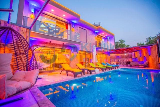 Akşam ışıklandırmalı, havuz kenarında şezlongların bulunduğu modern bir villa dış cephesi.