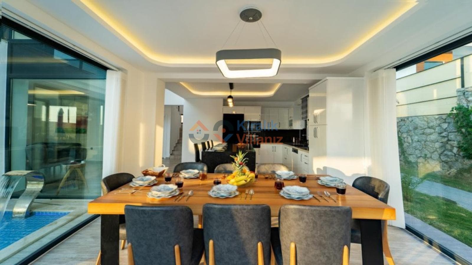 Ahşap yemek masası, gri sandalyeler ve modern aydınlatmalı açık mutfaklı yemek alanı.