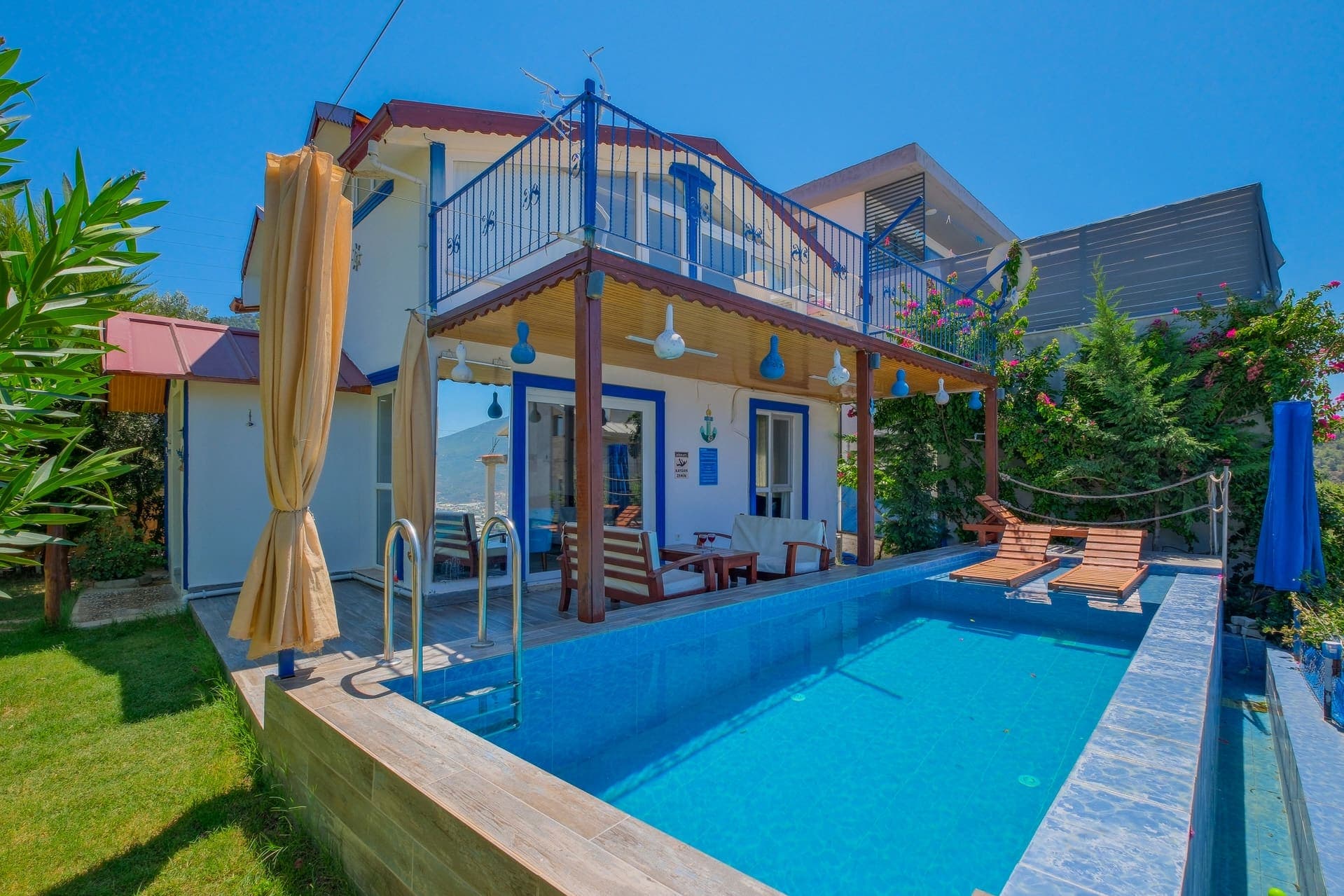 Önünde küçük bir havuz ve ahşap şezlonglar bulunan, balkonlu ve mavi detaylı villa.