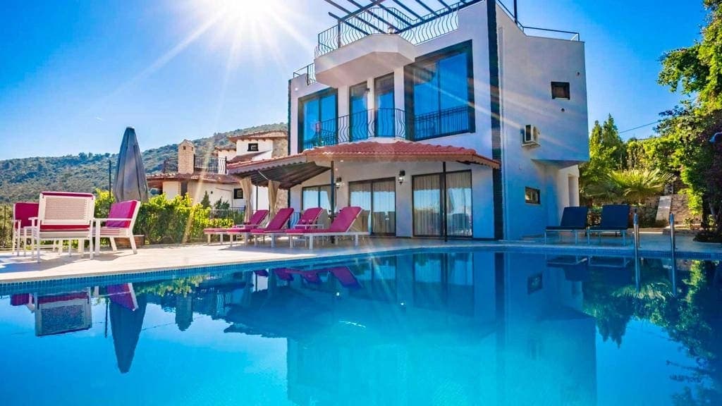 Güneşlenme terası, şezlonglar ve mavi havuzu olan beyaz dış cepheli iki katlı villa.