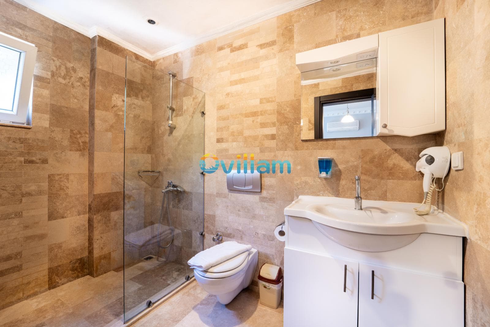 Bej tonlarında fayans kaplı, cam duşakabinli ve beyaz lavabolu banyo.