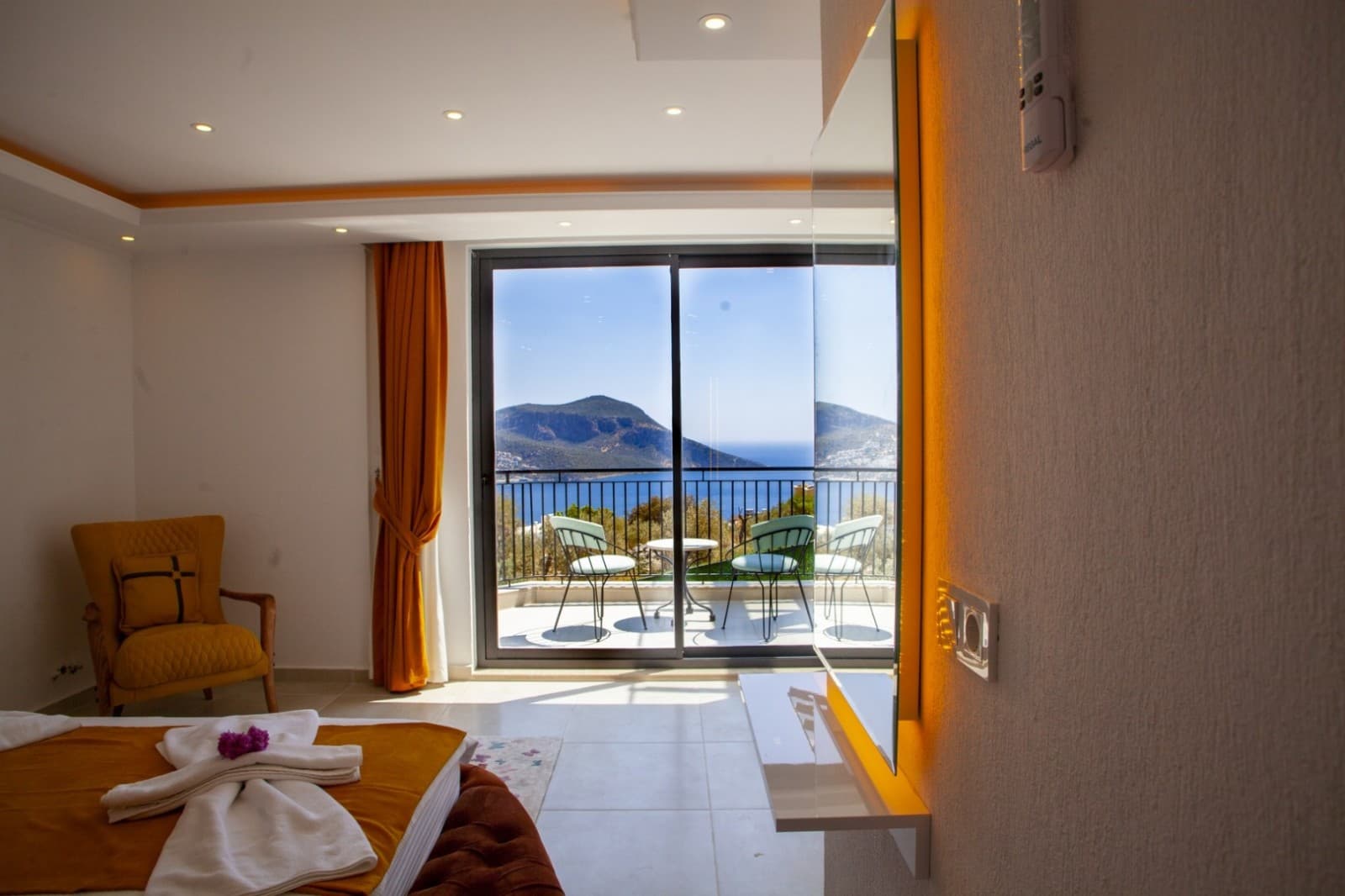Balkon kapısından deniz manzarası görünen, sarı detaylı yatak odası.