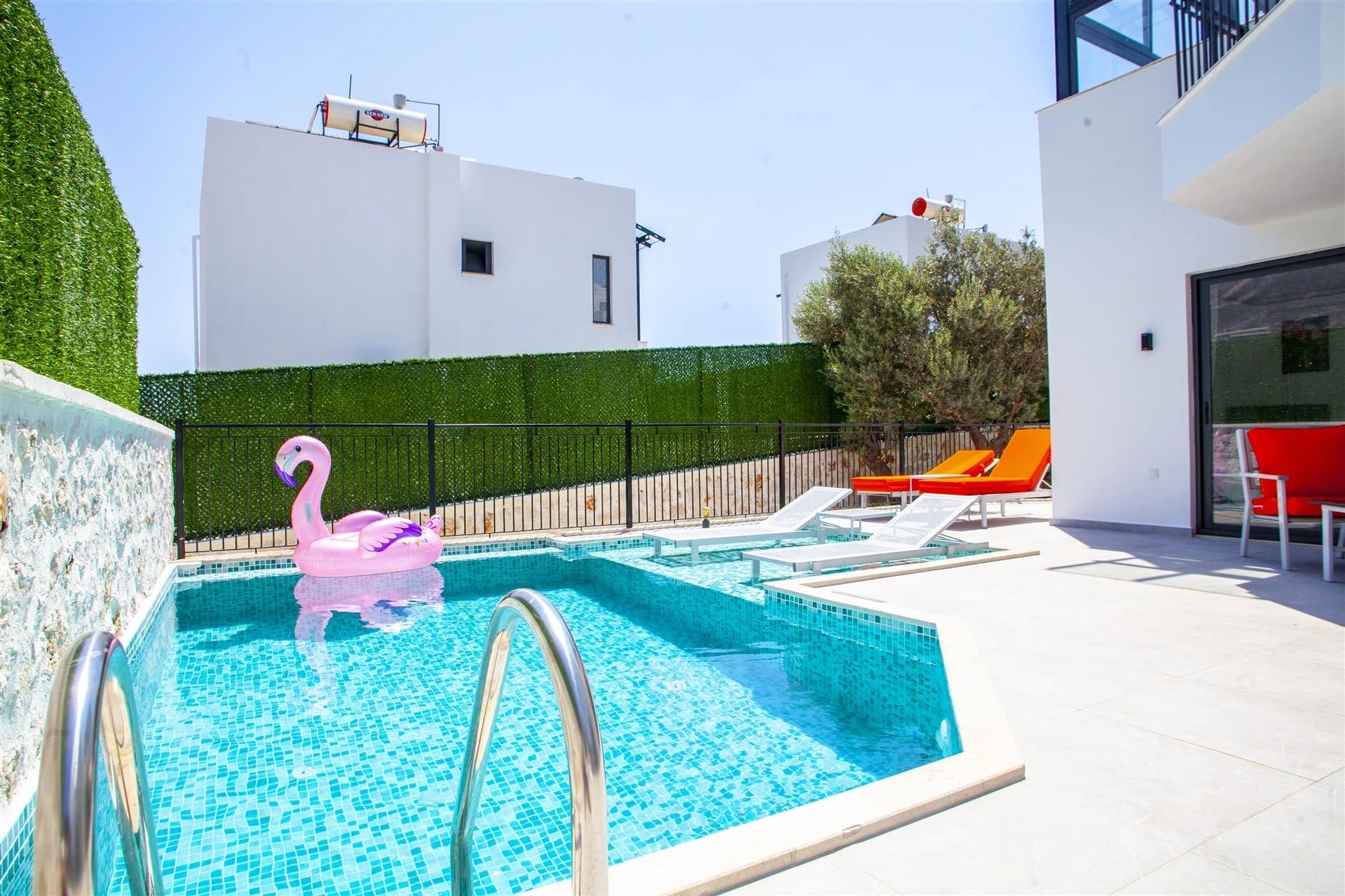 Pembe flamingo deniz yatağı bulunan havuzun kenarında turuncu şezlonglar ve beyaz villa yer alır.