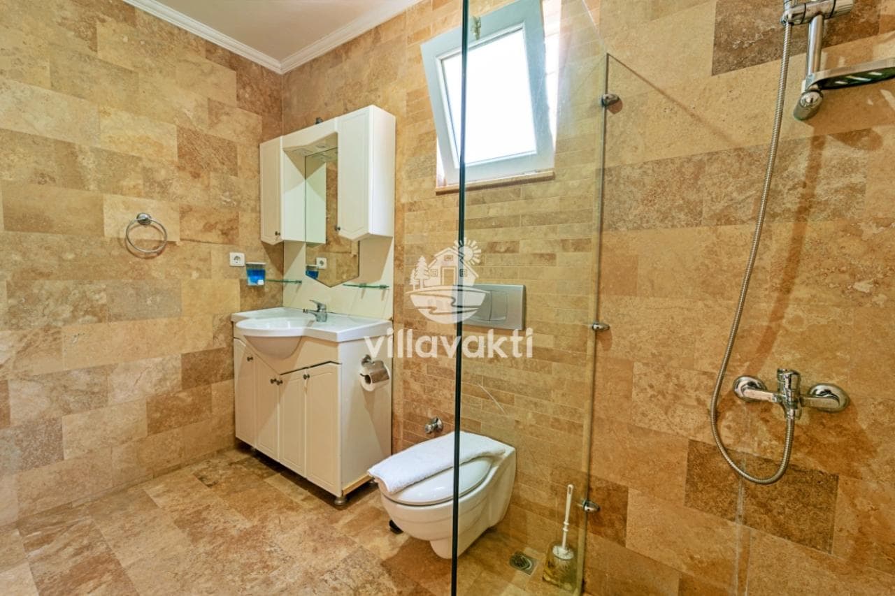 Bej renkli taş kaplamalı banyoda beyaz lavabo, klozet ve cam bölmeli duş alanı bulunur.