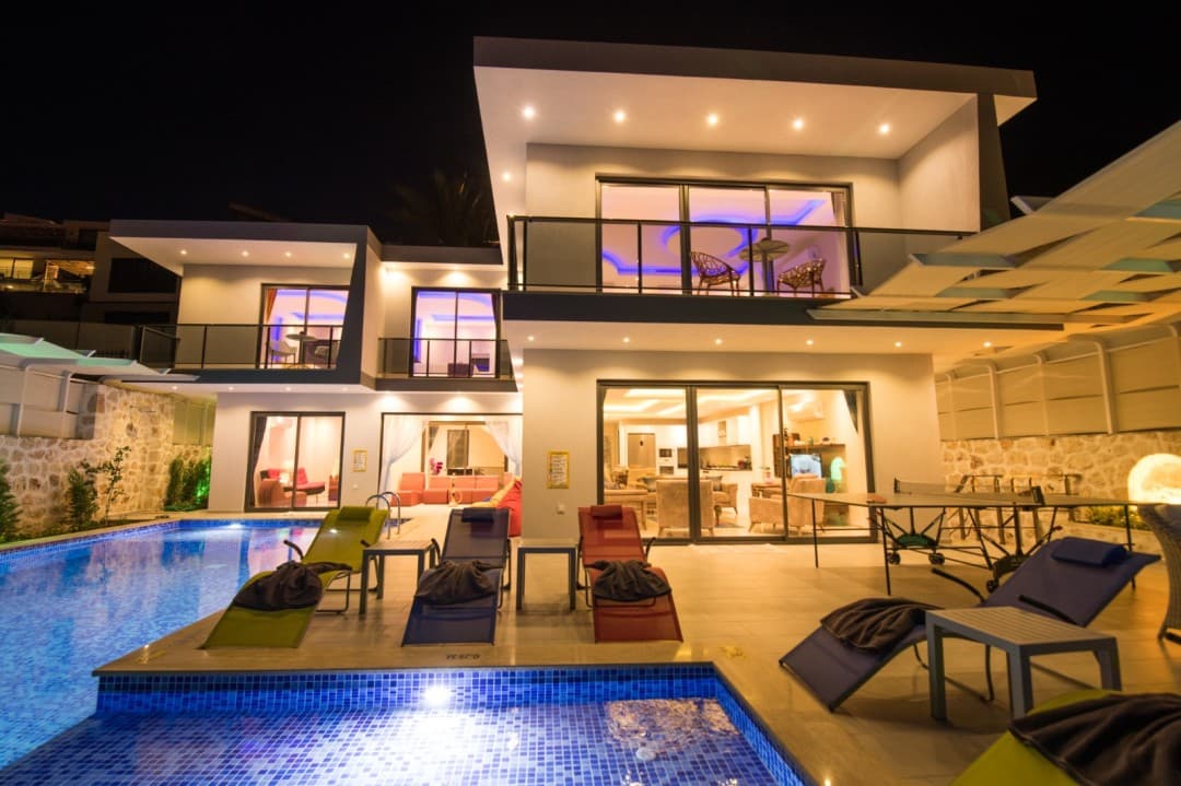 Gece aydınlatmalı, havuz başında şezlonglar ve masa tenisi bulunan iki katlı modern villa.