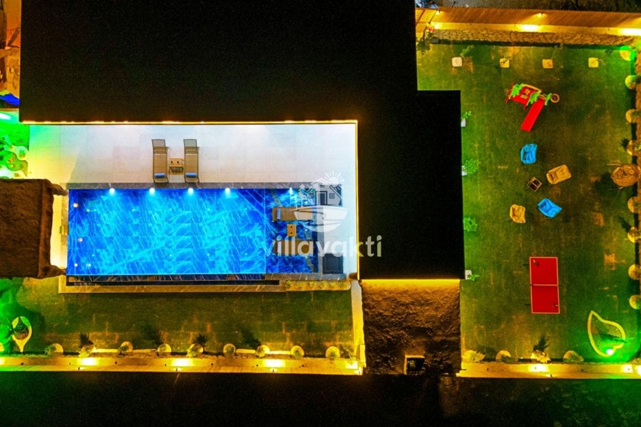 Gece aydınlatmalı yüzme havuzu ve çim bahçesi bulunan villanın kuş bakışı görünümü.