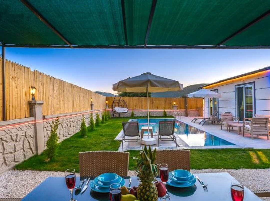 Ön planda yemek masası ve arkasında havuzlu villa bahçesi görünüyor.