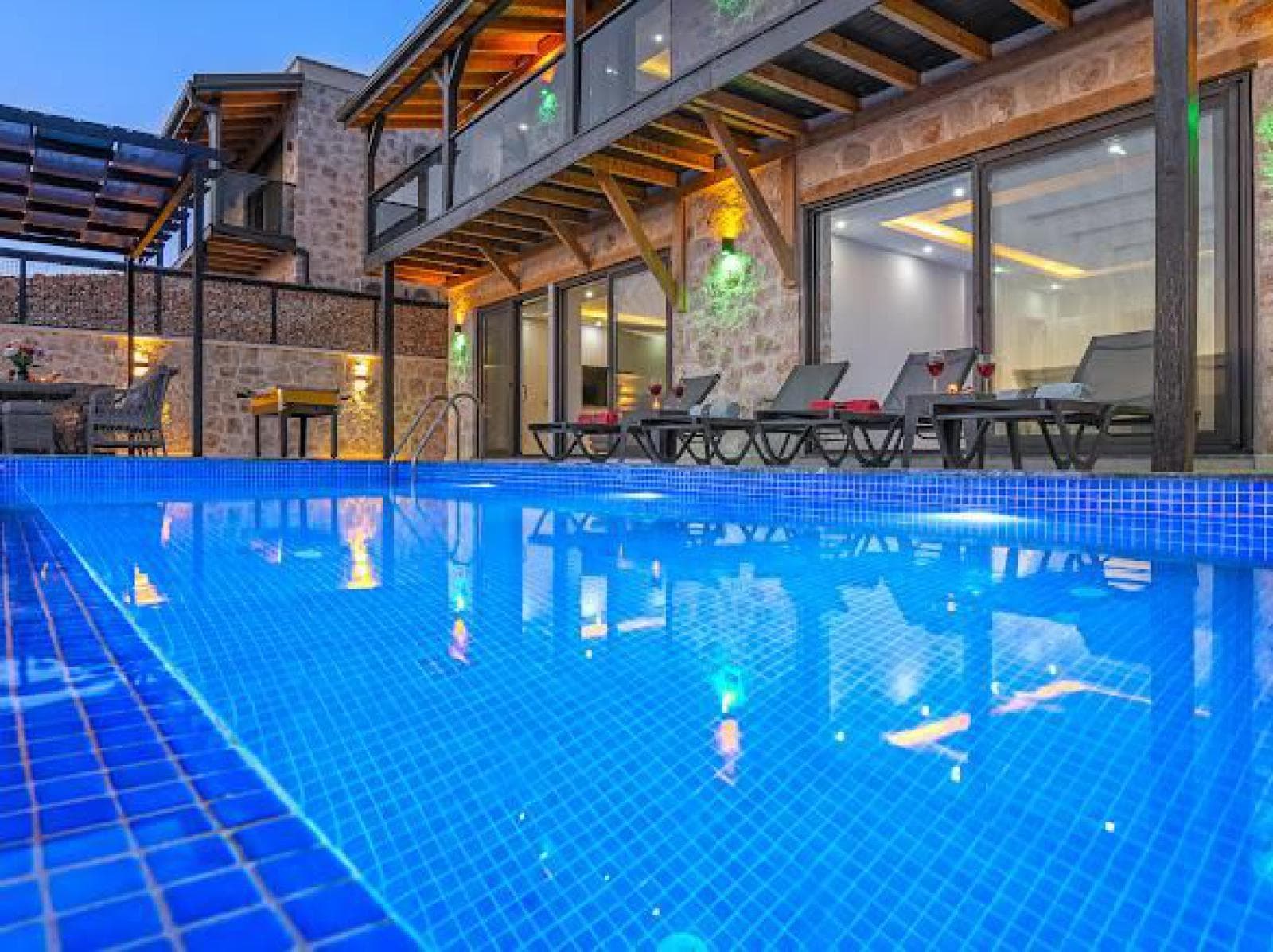 Akşam ışıklandırmalı havuz kenarında şezlonglar ve taş duvarlı villa cephesi.