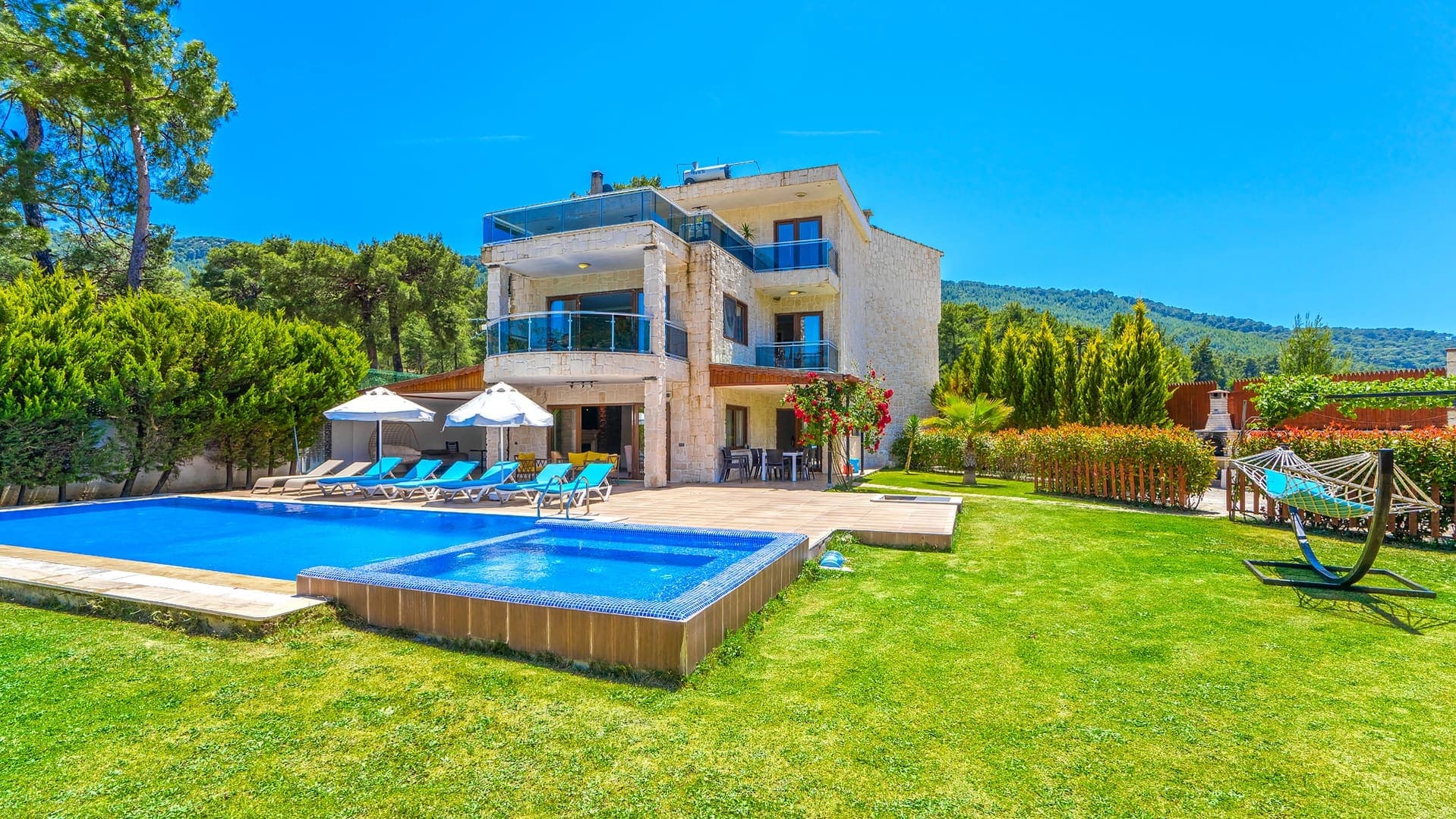 Yeşil ağaçlarla çevrili bahçesinde havuz, şezlonglar ve hamak bulunan üç katlı taş villa.