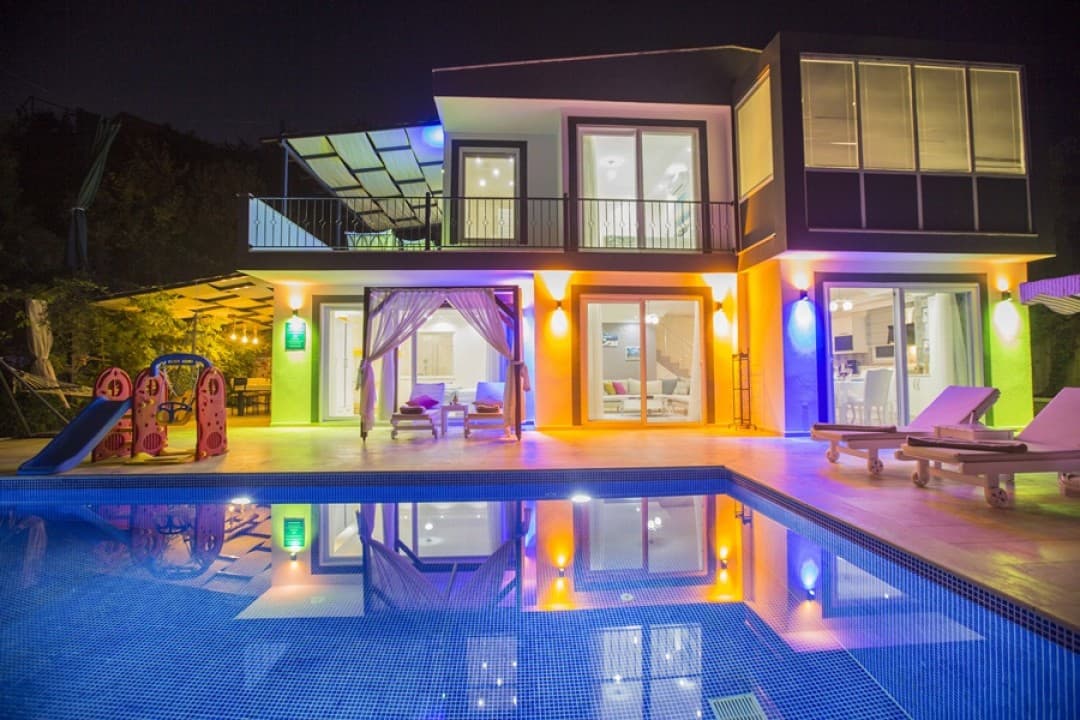 Gece aydınlatmalı, havuzlu, çocuk oyun parkı ve şezlongları bulunan iki katlı modern villa.