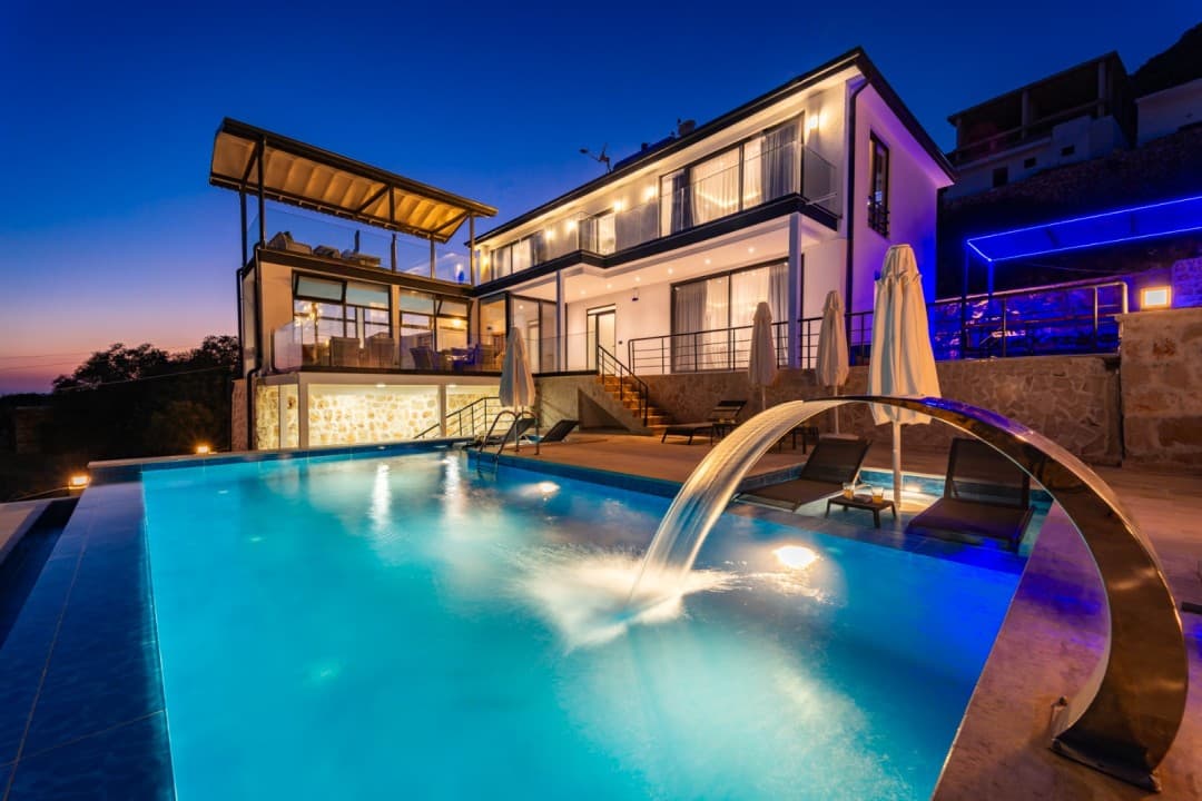 Akşam vakti aydınlatılmış, şelale özellikli havuzu ve güneşlenme şezlongları bulunan iki katlı modern villa.
