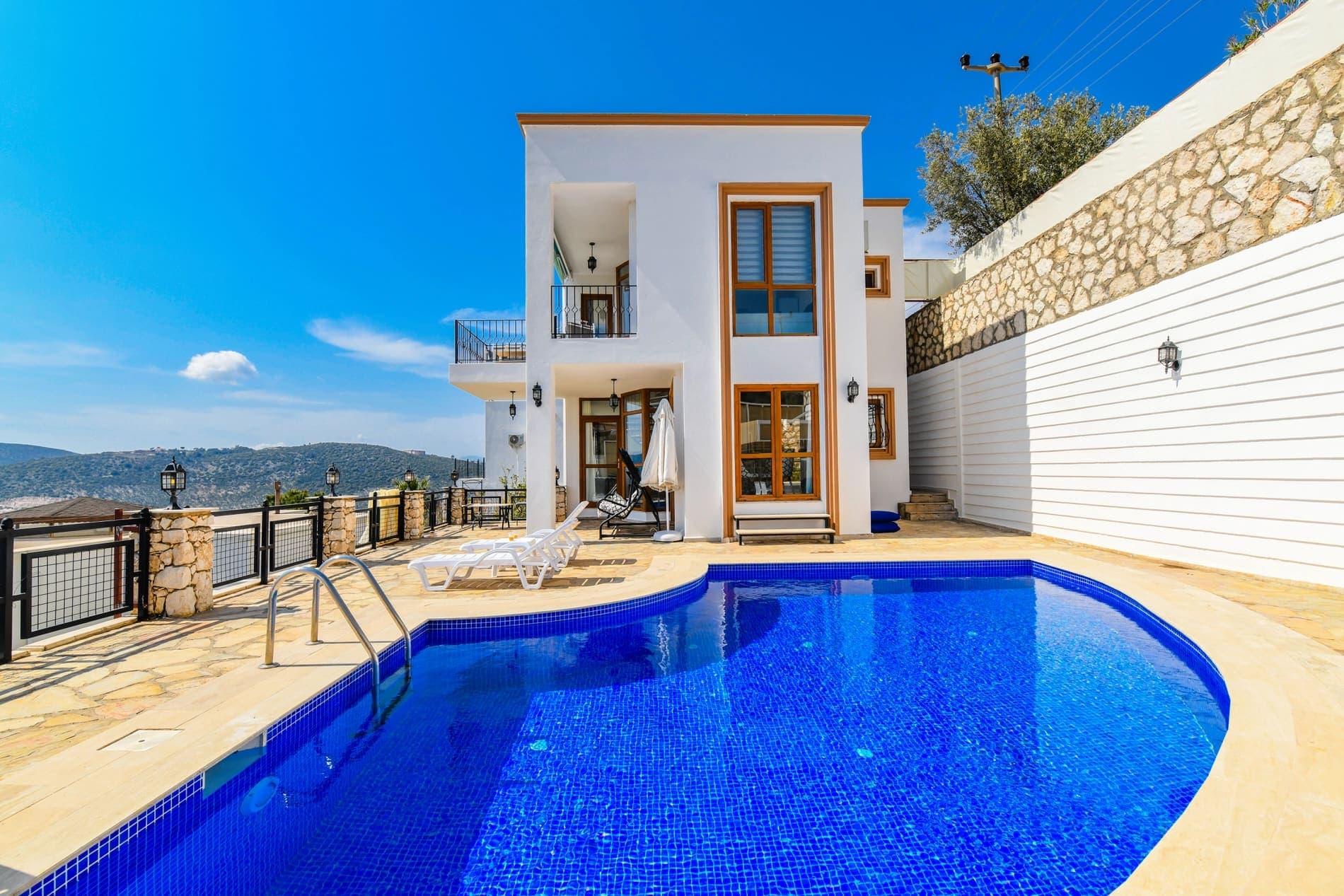 Açık mavi havuzu, şezlongları ve beyaz dış cephesi olan iki katlı villa.