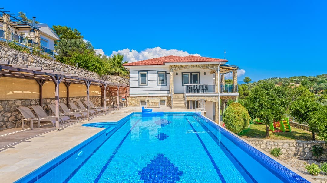 Ön planda uzun mavi havuz, yanında şezlonglar ve arka planda iki katlı villa yer alıyor.