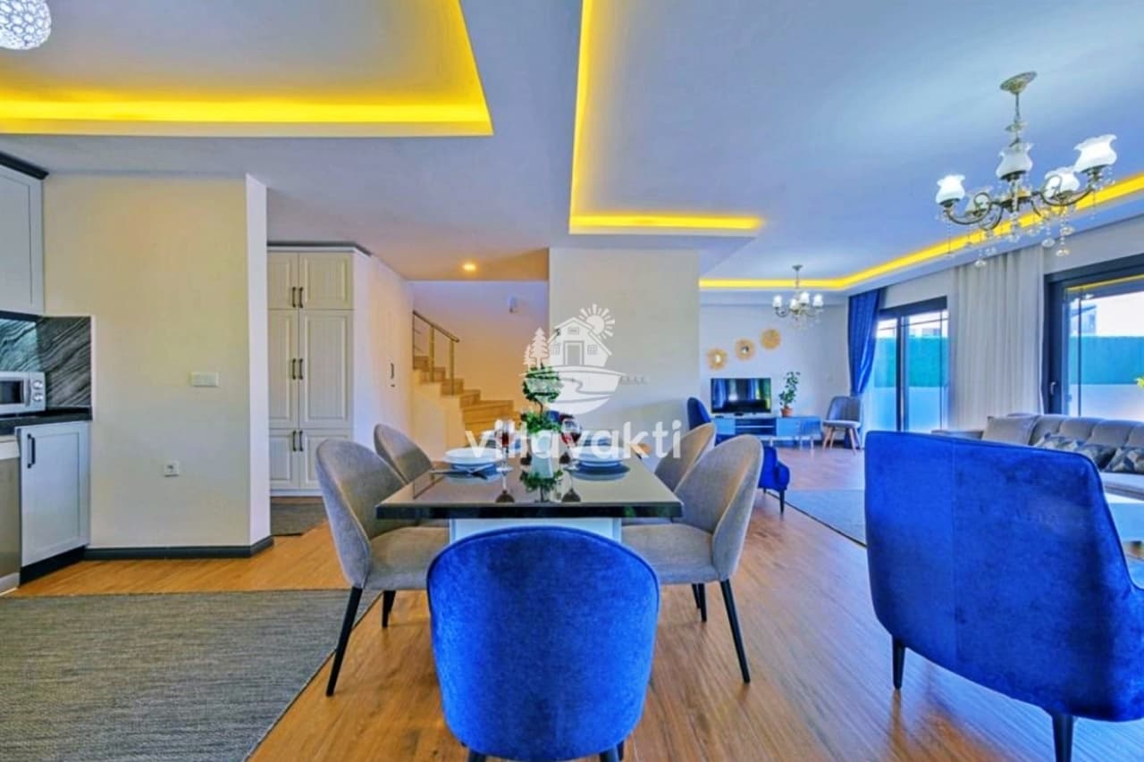 Açık plan salon ve yemek masası, mavi koltuklar ve sarı tavan aydınlatması.