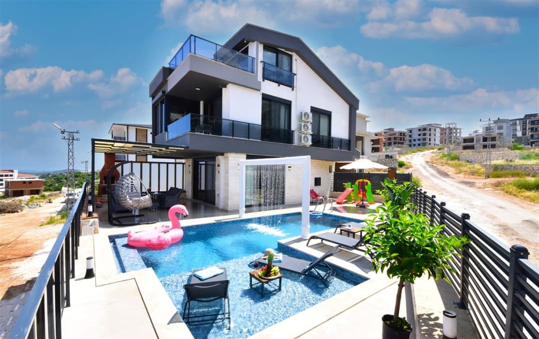 Önünde havuz, şezlonglar, pembe flamingo deniz yatağı ve çocuk oyun alanı bulunan modern üç katlı villa.