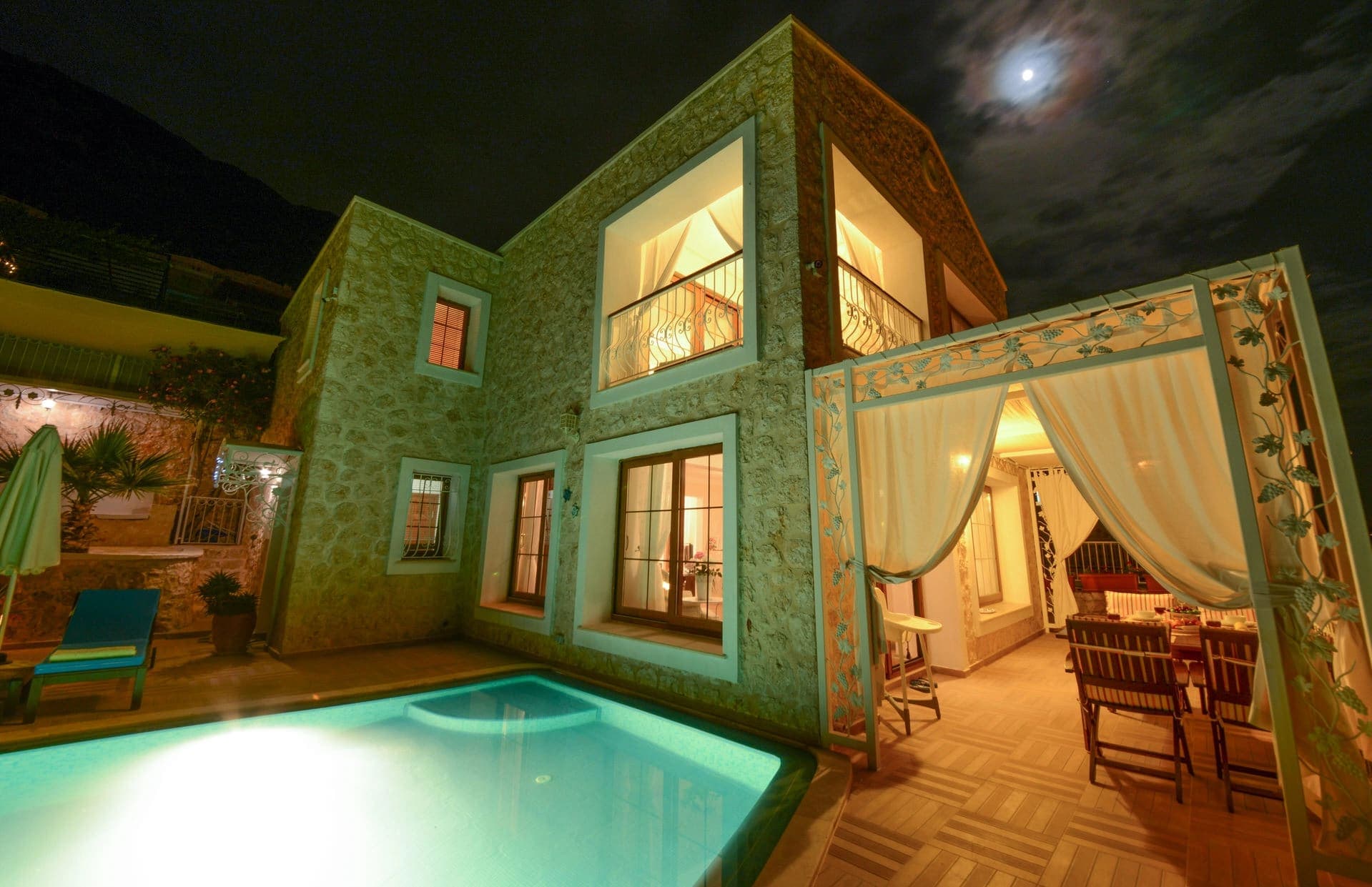Gece ışıklandırılmış taş villanın bahçesindeki yüzme havuzu ve perdeli çardak alanı.