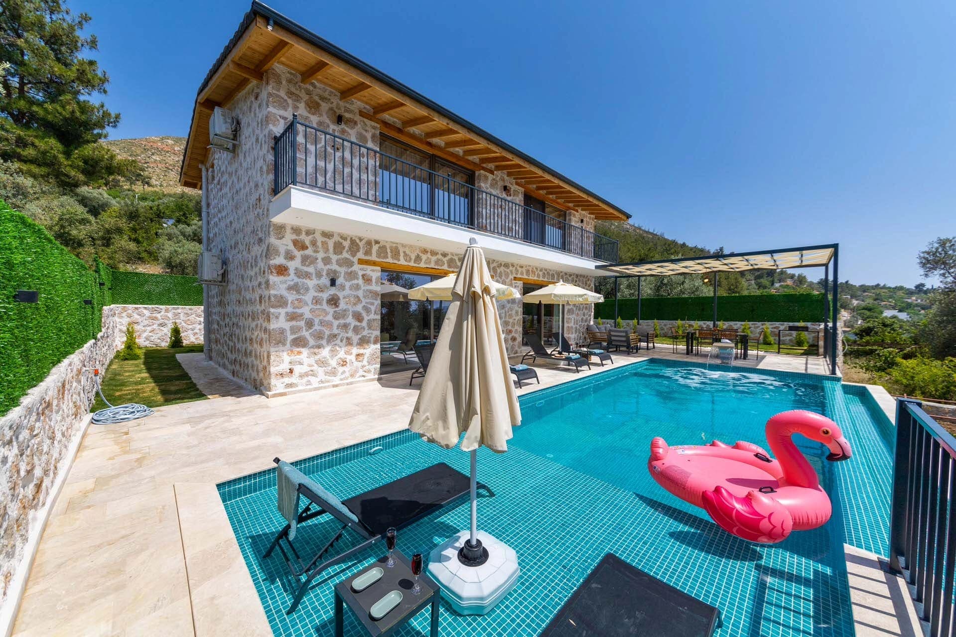 Taş duvarlı iki katlı villa, önünde havuz, pembe flamingo deniz yatağı ve şezlonglar.