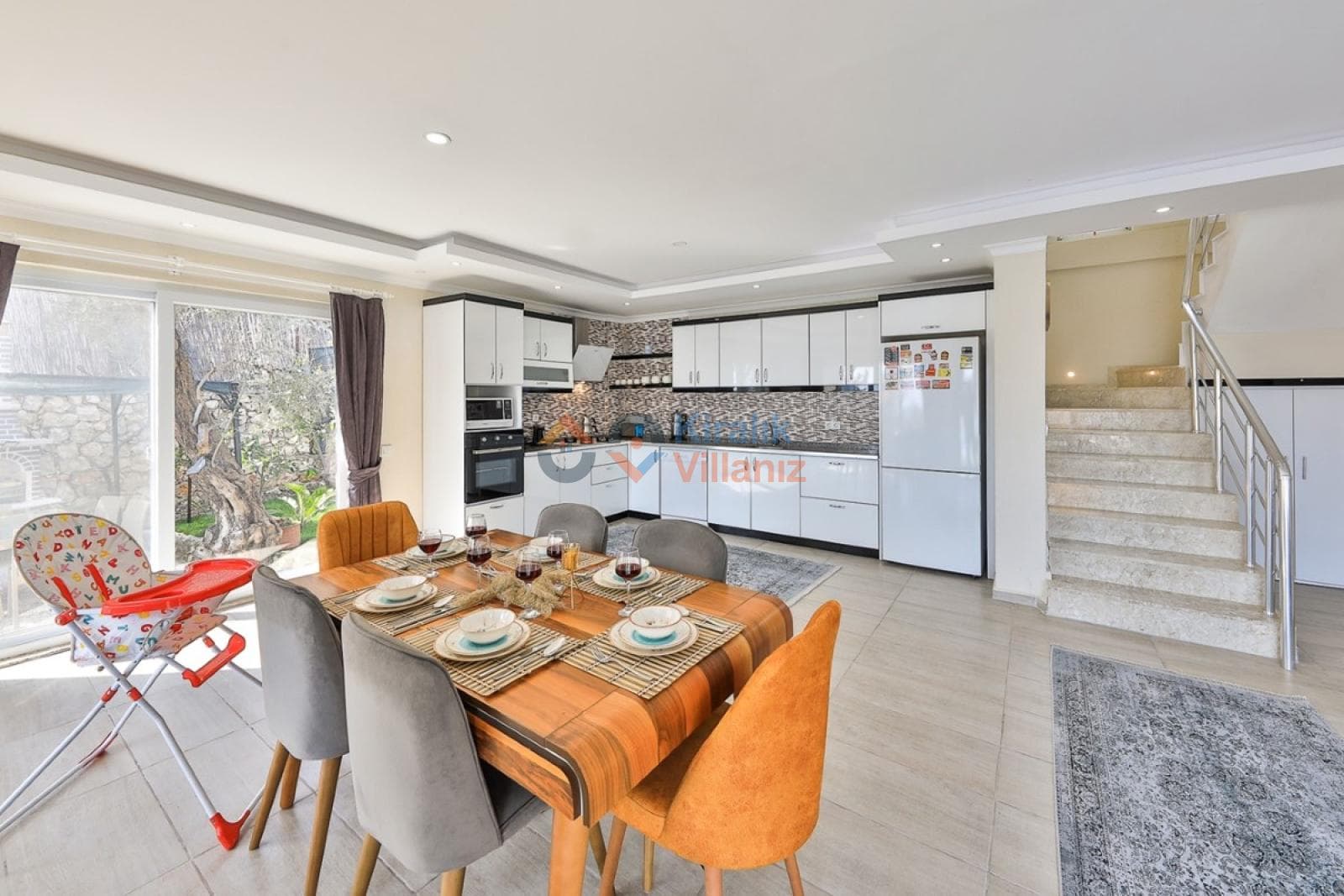 Beyaz dolaplı modern mutfak, ahşap yemek masası, turuncu sandalyeler ve üst kata çıkan merdivenler.