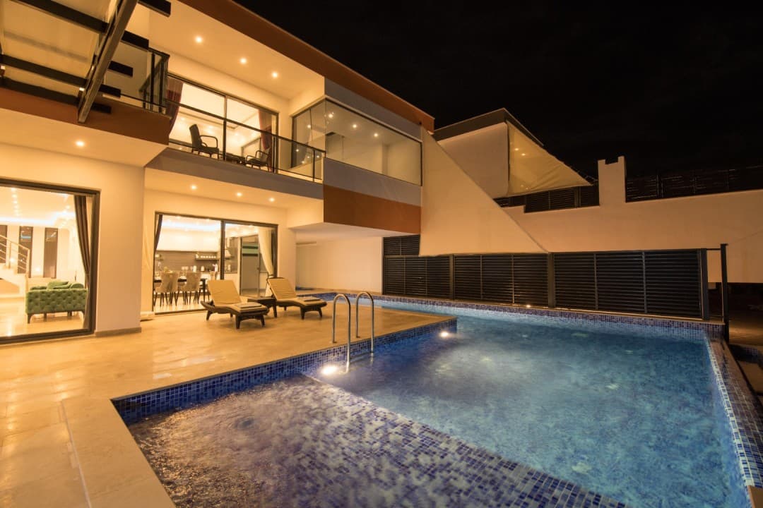 Gece aydınlatmalı havuzu, şezlongları ve balkonlu modern mimarisiyle iki katlı villa dış cephesi.