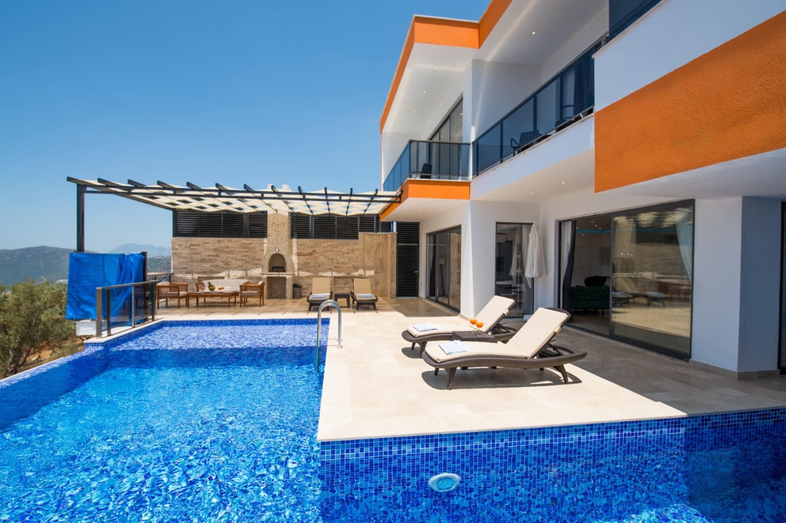 İki katlı villa önünde şezlonglu havuz ve ahşap oturma grubu.