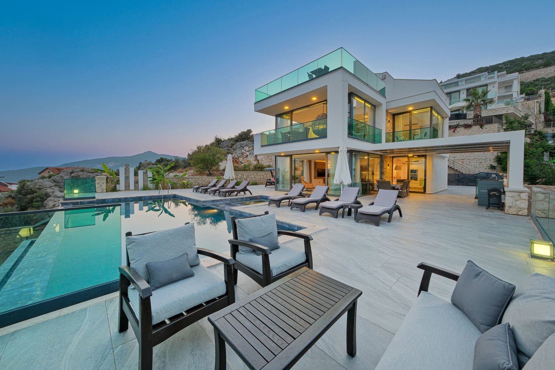 Modern mimarili beyaz villa, geniş teras, gri oturma grubu ve yüzme havuzu.