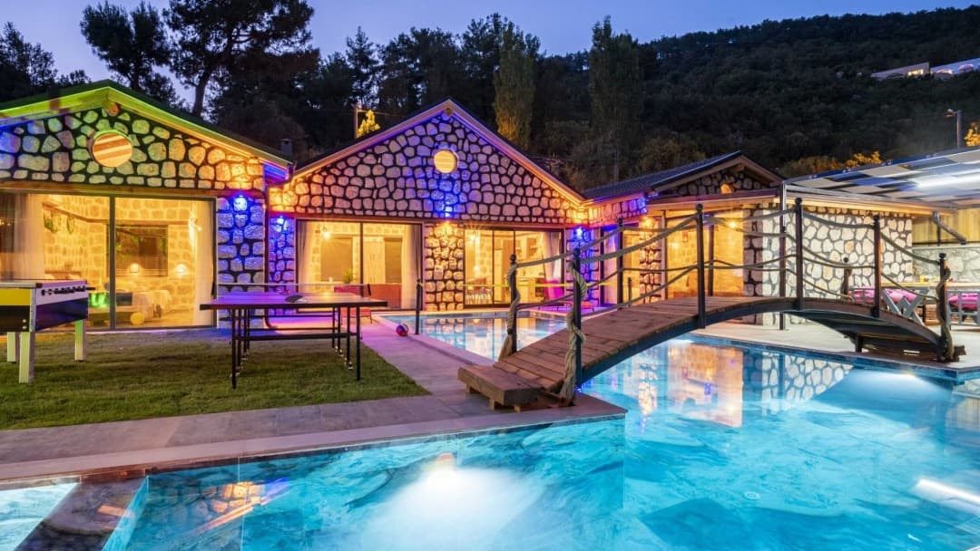 Gece aydınlatmalı taş duvarlı villa, önünde havuz, ahşap köprü, masa tenisi ve langırt masası bulunuyor.