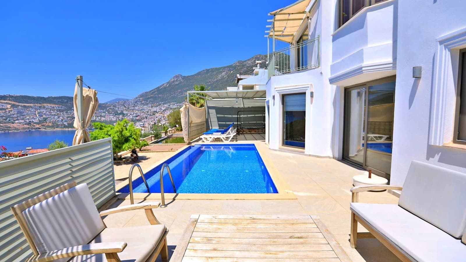 Deniz manzaralı havuz kenarında beyaz şezlonglar ve oturma grubu bulunan villa cephesi.
