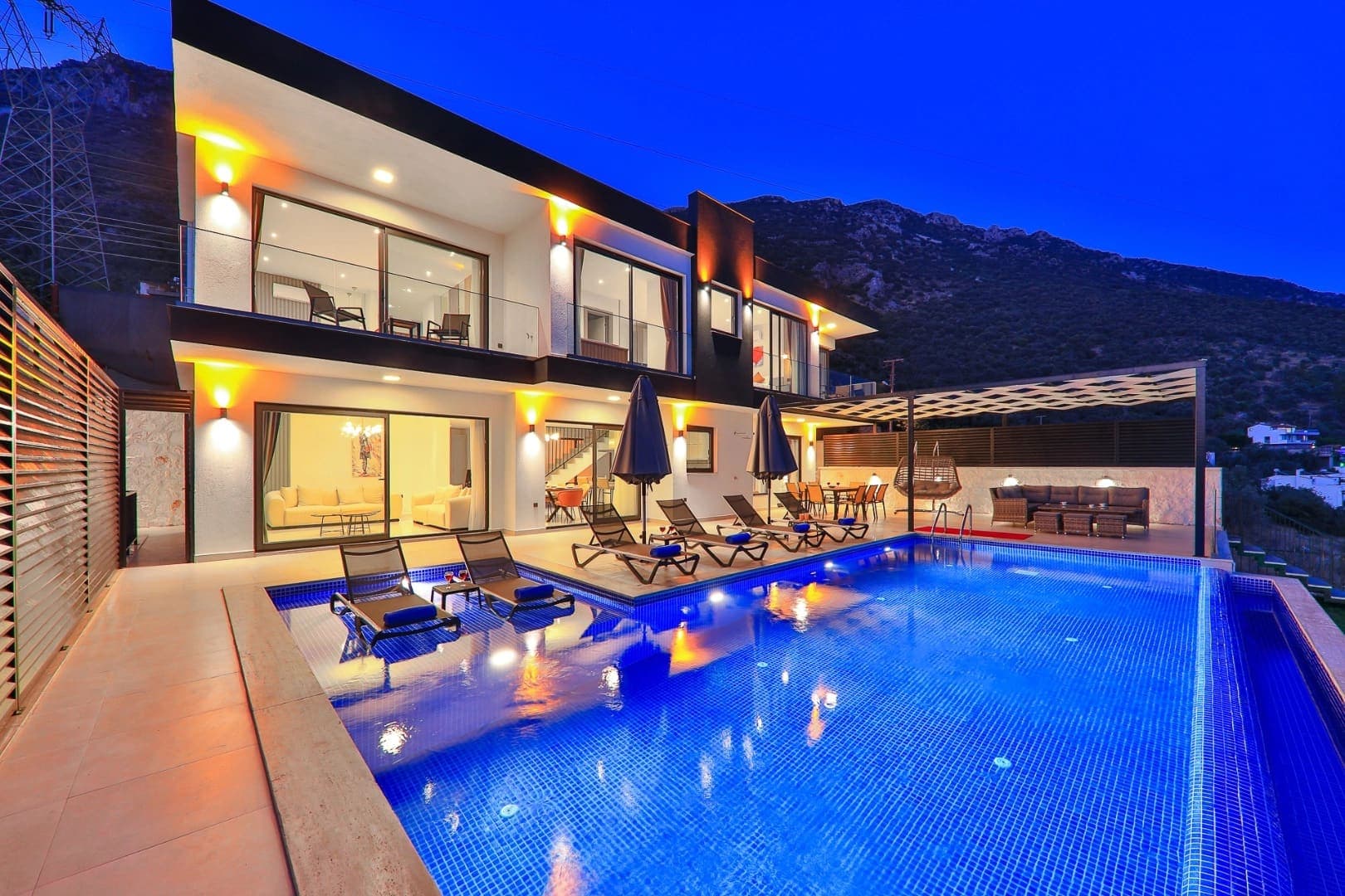 Gece ışıklandırmalı havuz ve önünde şezlonglar bulunan modern villa dış cephesi.