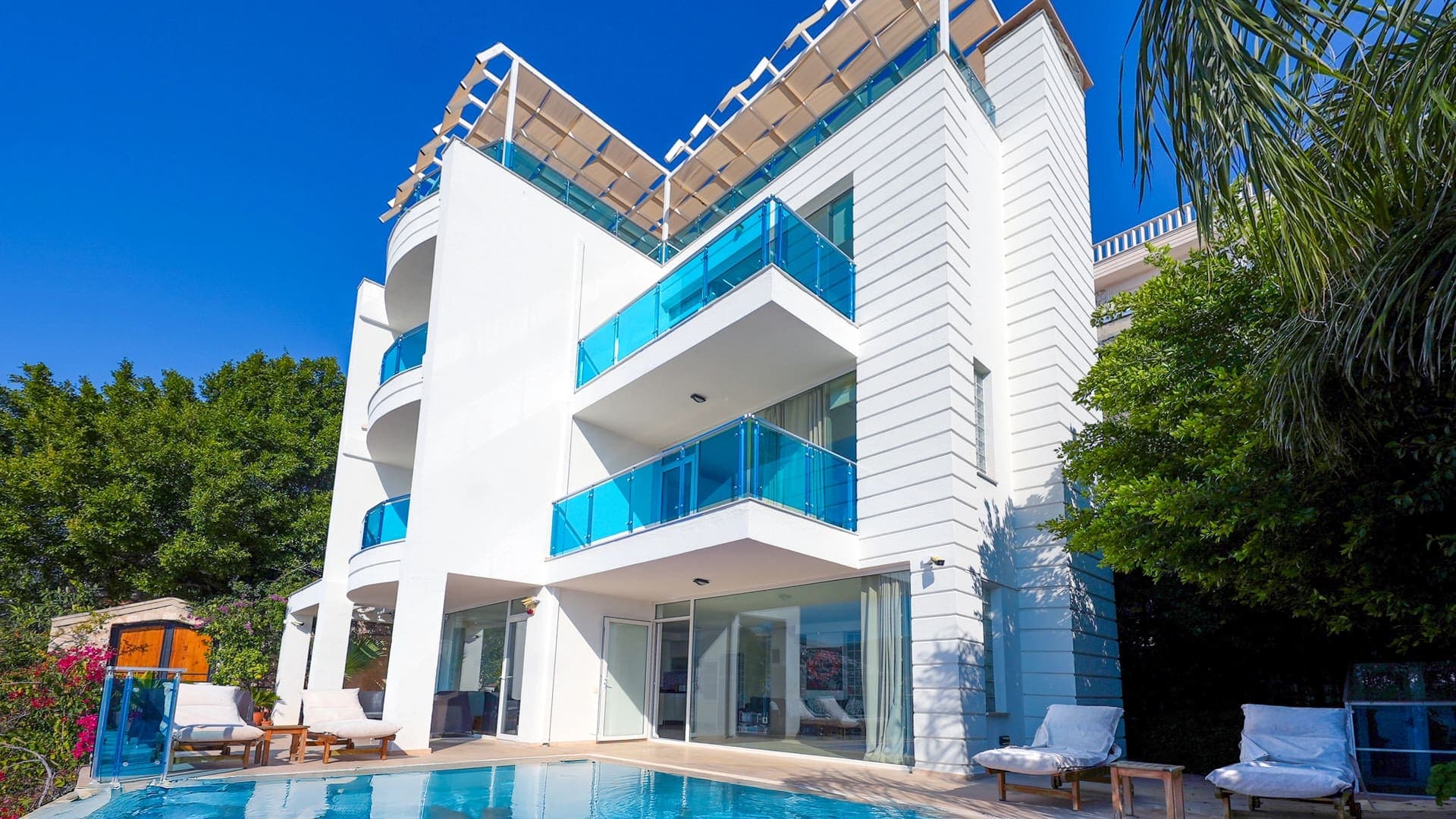 Açık mavi havuz kenarında şezlonglar bulunan beyaz villa dış cephesi.