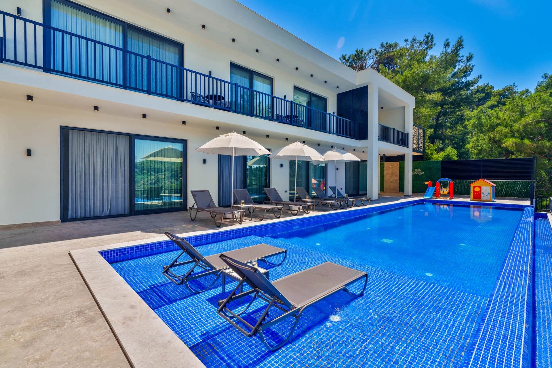 Geniş yüzme havuzu ve şezlongları bulunan beyaz iki katlı villa.
