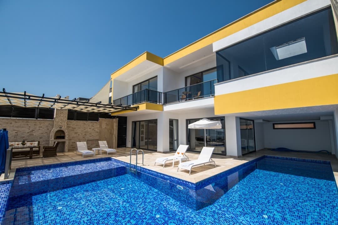 Mavi mozaik kaplı havuzu, şezlongları ve sarı beyaz dış cephesi olan iki katlı modern bir villa.