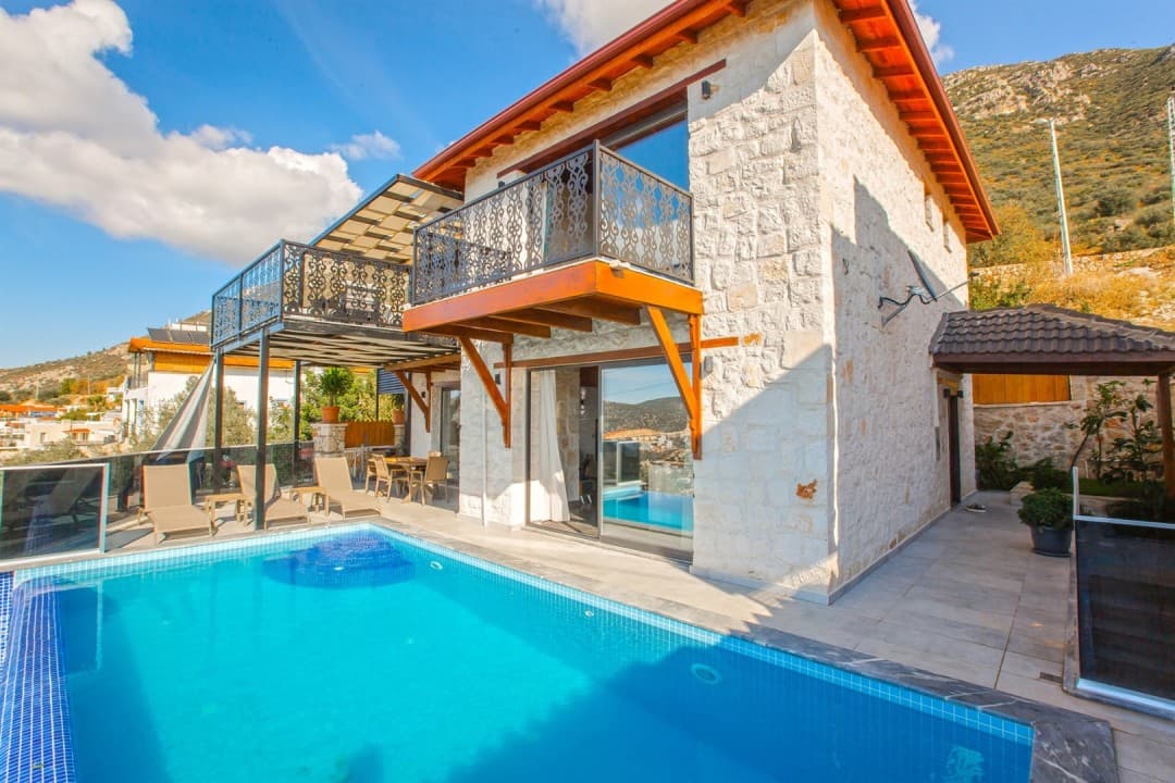 Taş duvarlı, balkonlu ve önünde özel yüzme havuzu ile şezlonglar bulunan iki katlı villa.