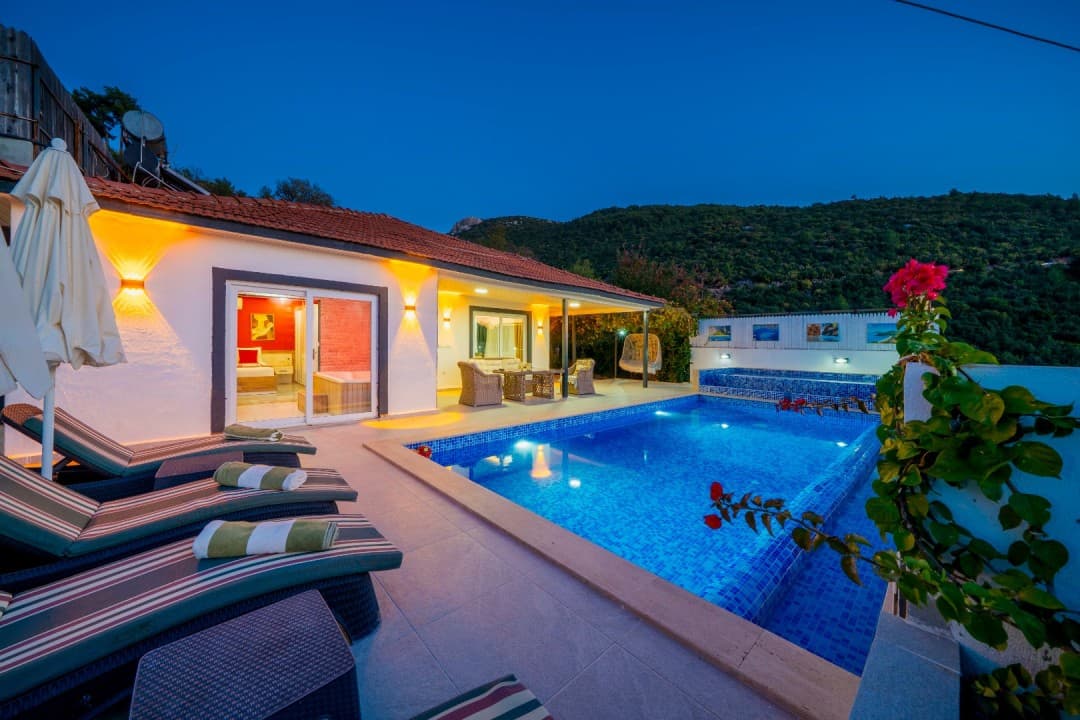 Akşam vakti havuz başı, şezlonglar ve aydınlatılmış villa dış görünümü.